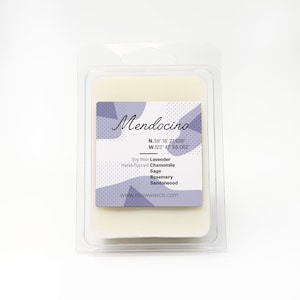May include: A white soy wax candle in a clear plastic container with a purple and white label that reads "Mendocino" with coordinates and a list of scents: Lavender, Chamomile, Sage, Rosemary, Sandalwood. The website address is www.neswwaxco.com.