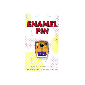 May include: An enamel pin featuring a retro-style instant camera in yellow, orange, and purple. The pin includes a photo of a mountain landscape. The words "ENAMEL PIN" are in red and black. The website address is also visible.