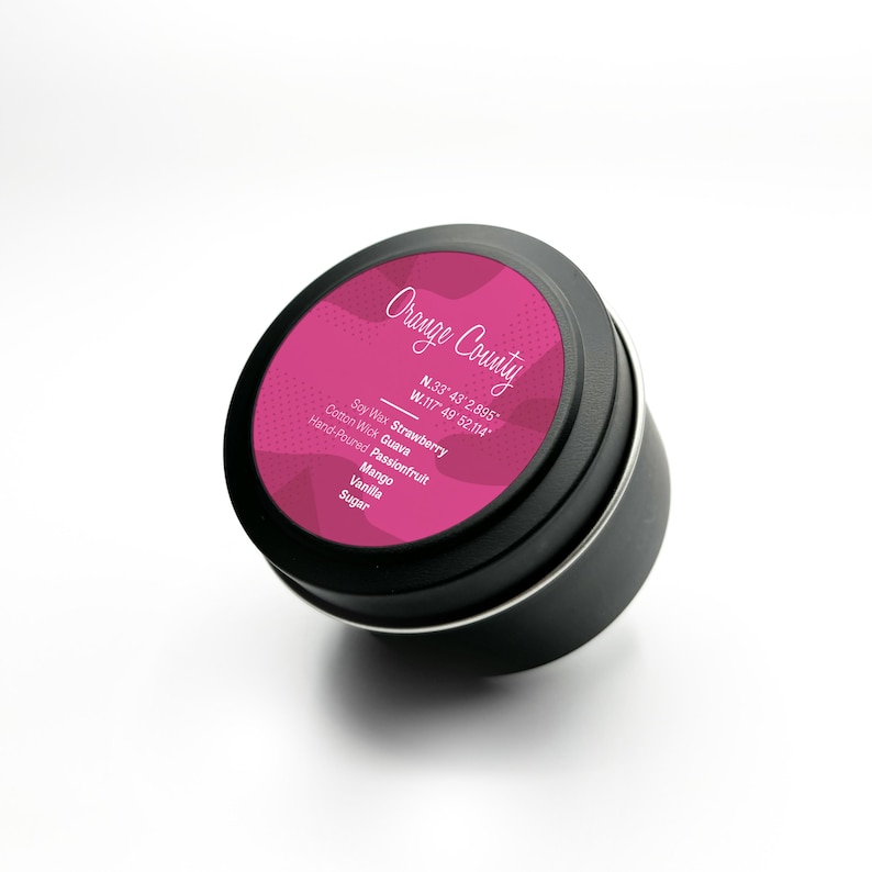 Puede incluir: Una vela de metal negro en una lata con una etiqueta rosa que dice "Orange County" y las coordenadas "N.33&deg; 43' 2.895' W.117&deg; 49' 52.114'". La etiqueta tambi&eacute;n indica el aroma "Fresa" e incluye los ingredientes: Cera de soja, Guayaba, Maracuy&aacute;, Mango, Vainilla, Az&uacute;car.