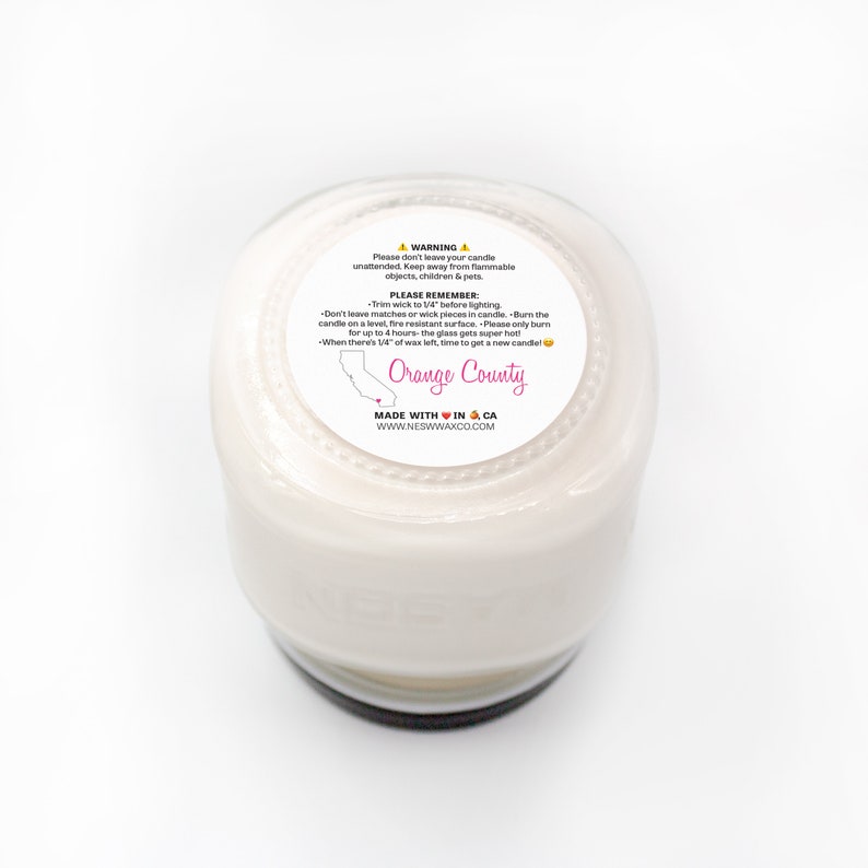 Puede incluir: Un tarro de vela blanco con una tapa negra y una etiqueta que dice "Orange County" con un contorno de California y un coraz&oacute;n. La etiqueta tambi&eacute;n incluye el texto "Made with Love in CA" y "www.nesnwaxco.com".