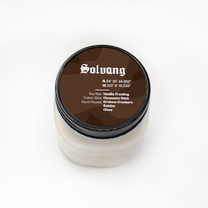 May include: A brown glass jar candle with a black lid. The lid has a brown label with white text that reads "Solvang", "N.34° 35' 44.952", "W.120° 8' 15.533", "Soy Wax Vanilla Frosting", "Cotton Wick Cinnamon Stick", "Hand-Poured Graham Crackers", "Raisins", and "Clove".
