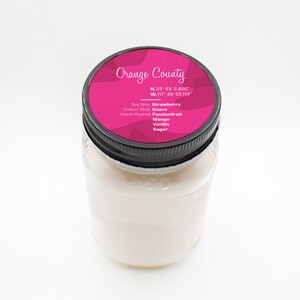 Puede incluir: Una vela de vidrio en un frasco con una tapa negra. La tapa tiene una etiqueta rosa con el texto "Orange County" y coordenadas. La etiqueta tambi&eacute;n enumera las fragancias: Fresa, Guayaba, Maracuy&aacute;, Mango, Vainilla y Az&uacute;car. La vela est&aacute; hecha a mano y hecha con cera de soya y una mecha de algod&oacute;n.