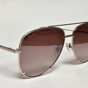 Puede incluir: Gafas de sol de aviador doradas con lentes marrones degradadas. La marca "DIFF" es visible en la montura y la patilla. Las gafas tienen un doble puente y almohadillas nasales. Las lentes pasan de un marr&oacute;n m&aacute;s oscuro en la parte superior a un tono m&aacute;s claro en la inferior.