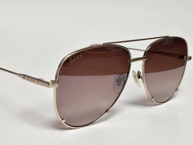Puede incluir: Gafas de sol de aviador doradas con lentes marrones degradadas. La marca "DIFF" es visible en la montura y la patilla. Las gafas tienen un doble puente y almohadillas nasales. Las lentes pasan de un marr&oacute;n m&aacute;s oscuro en la parte superior a un tono m&aacute;s claro en la inferior.