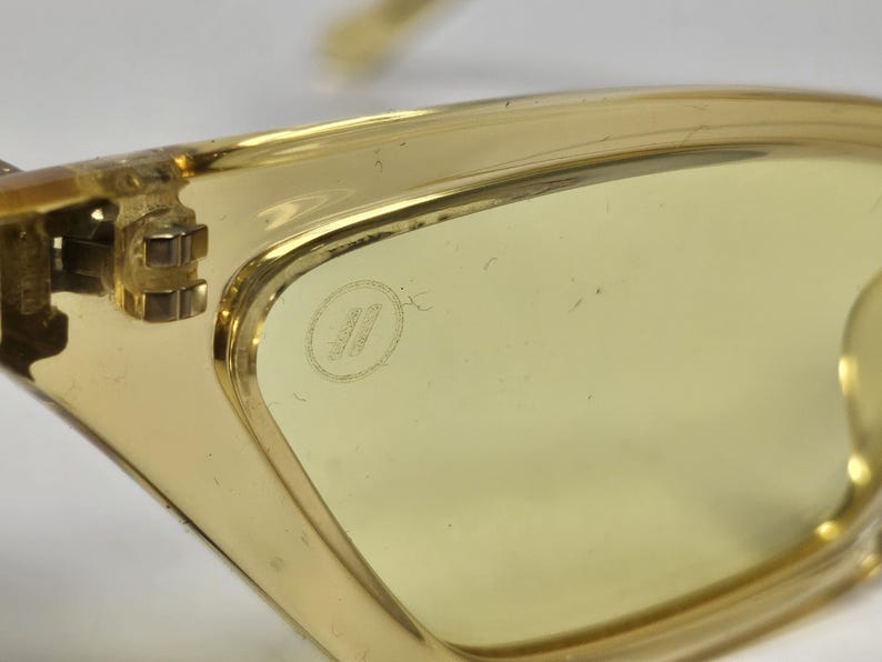 K&ouml;nnte beinhalten: Durchscheinende, gelb get&ouml;nte Brille mit einem klaren Kunststoffrahmen. Das Glas zeigt ein kleines, kreisf&ouml;rmiges, eingraviertes Logo in der N&auml;he des Scharniers. Der Metallscharnier-Mechanismus ist sichtbar und verbindet den Rahmen mit dem B&uuml;gel.