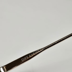 Puede incluir: Primer plano de una parte de gafas. La montura met&aacute;lica plateada tiene el texto "DIFF EYEWEAR" grabado en la varilla. El fondo es blanco liso.