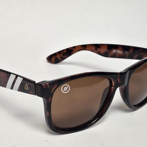 May include: Tortoiseshell sunglasses with brown lenses. The left temple features two white stripes and a small gold triangle logo. The right lens has a white circular logo with two vertical lines. The inner temple is marked with M Class and CE.