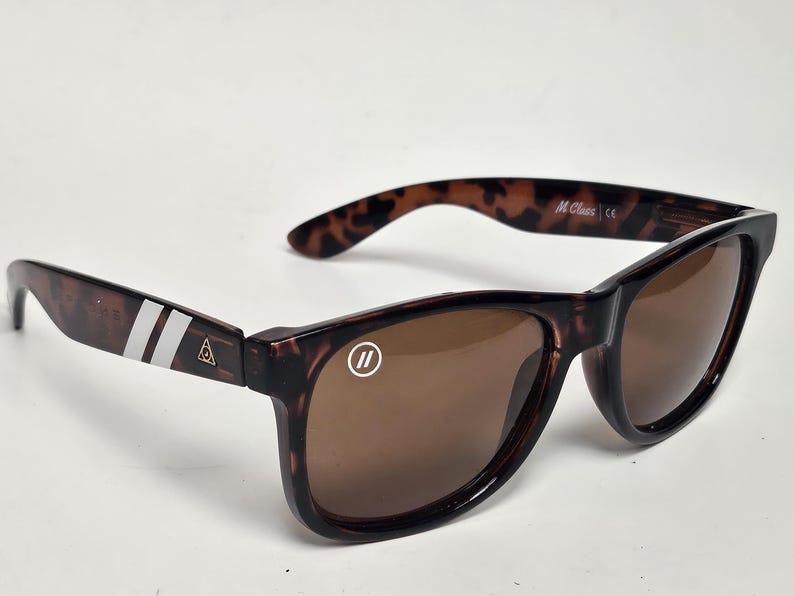 May include: Tortoiseshell sunglasses with brown lenses. The left temple features two white stripes and a small gold triangle logo. The right lens has a white circular logo with two vertical lines. The inner temple is marked with M Class and CE.