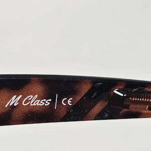 May include: The inner temple of a pair of eyeglasses featuring a brown and black tortoiseshell pattern. The text M Class and the CE mark are printed in white on the inner surface of the frame arm, which also shows a visible metal hinge mechanism.