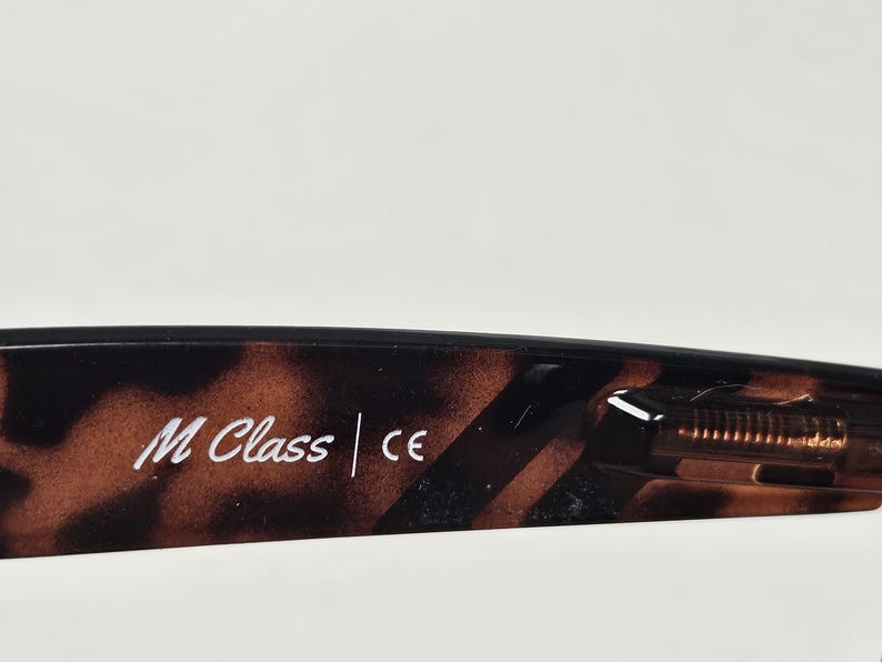 May include: The inner temple of a pair of eyeglasses featuring a brown and black tortoiseshell pattern. The text M Class and the CE mark are printed in white on the inner surface of the frame arm, which also shows a visible metal hinge mechanism.