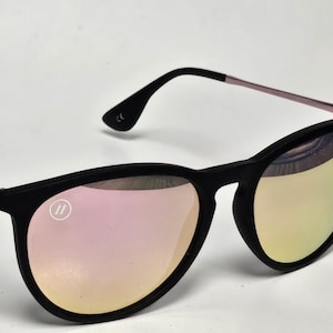 May include: Sunglasses with a matte black frame and reflective pink-tinted lenses. The temples are thin and metallic pink. A small circular logo with two vertical lines is visible on the upper left lens, and the CE mark is printed on the inner temple.