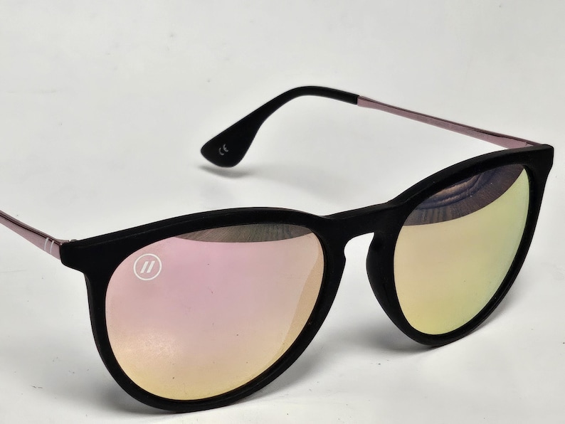 May include: Sunglasses with a matte black frame and reflective pink-tinted lenses. The temples are thin and metallic pink. A small circular logo with two vertical lines is visible on the upper left lens, and the CE mark is printed on the inner temple.