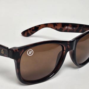 May include: Tortoiseshell sunglasses with brown lenses. The left lens features a white circular logo with two vertical lines. The temples display two white stripes and a small gold triangular emblem. The inner temple is printed with M Class.