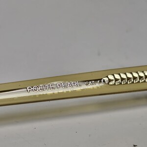 K&ouml;nnte beinhalten: Ein durchscheinender beigefarbener Brillenb&uuml;gel mit dem in Silber gedruckten Text POLITE PEARL CAT. 1. Der B&uuml;gel weist ein strukturiertes, sich wiederholendes blattartiges Muster auf, das entlang der Seite bis zum Ende des Rahmenteils verl&auml;uft.