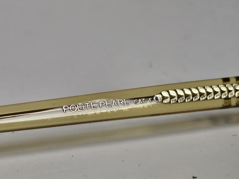 K&ouml;nnte beinhalten: Ein durchscheinender beigefarbener Brillenb&uuml;gel mit dem in Silber gedruckten Text POLITE PEARL CAT. 1. Der B&uuml;gel weist ein strukturiertes, sich wiederholendes blattartiges Muster auf, das entlang der Seite bis zum Ende des Rahmenteils verl&auml;uft.