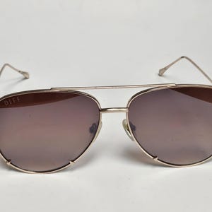 Puede incluir: Gafas de sol de aviador doradas con lentes tintadas marrones. Las gafas de sol tienen un doble puente y la marca "DIFF" es visible en la lente izquierda. Las monturas son de color dorado claro.