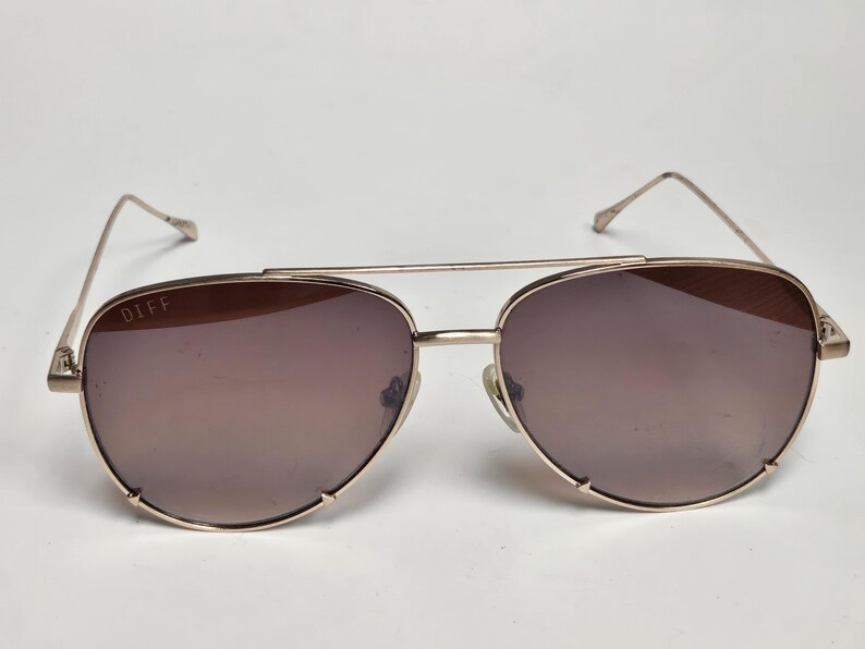 Puede incluir: Gafas de sol de aviador doradas con lentes tintadas marrones. Las gafas de sol tienen un doble puente y la marca "DIFF" es visible en la lente izquierda. Las monturas son de color dorado claro.