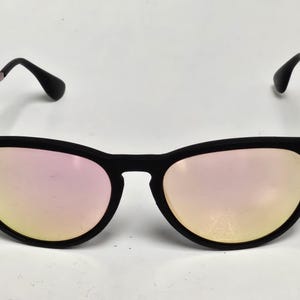 May include: Sunglasses with a matte black frame and reflective pink-tinted lenses. The thin metal temples are light pink, and the black temple tips are curved. A small circular logo is visible on the upper corner of the left lens.