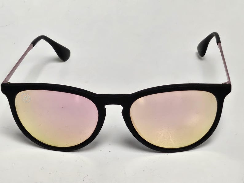 May include: Sunglasses with a matte black frame and reflective pink-tinted lenses. The thin metal temples are light pink, and the black temple tips are curved. A small circular logo is visible on the upper corner of the left lens.