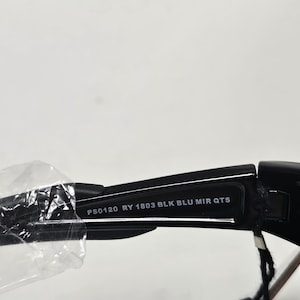 K&ouml;nnte beinhalten: Nahaufnahme einer schwarzen Sonnenbrille mit verspiegelten Gl&auml;sern. Der Text "PS0120 RY 1803 BLK BLU MIR QTS" ist auf dem B&uuml;gel der Sonnenbrille aufgedruckt. Die Sonnenbrille ist teilweise in Klarsichtfolie verpackt.