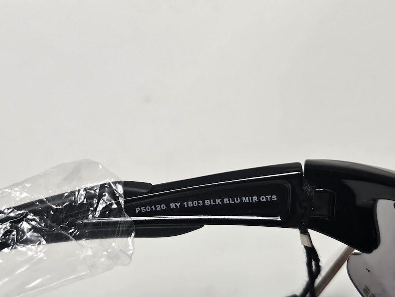 K&ouml;nnte beinhalten: Nahaufnahme einer schwarzen Sonnenbrille mit verspiegelten Gl&auml;sern. Der Text "PS0120 RY 1803 BLK BLU MIR QTS" ist auf dem B&uuml;gel der Sonnenbrille aufgedruckt. Die Sonnenbrille ist teilweise in Klarsichtfolie verpackt.