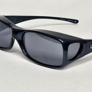 May include: Dark navy blue sunglasses with a wrap-around frame design and dark grey tinted lenses. The temples feature a silver-toned metal logo emblem with the letter J. The inner temple is printed with the text 61-19-128 ARIA 001.