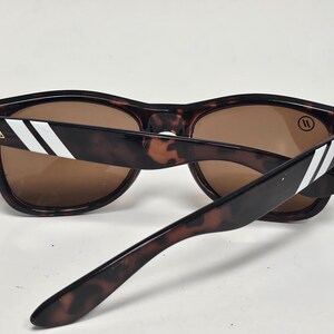 May include: Tortoiseshell sunglasses with brown lenses. The temples feature two white diagonal stripes and a small gold triangular logo near the hinges. A circular logo with two vertical lines is visible on the upper corner of the right lens.