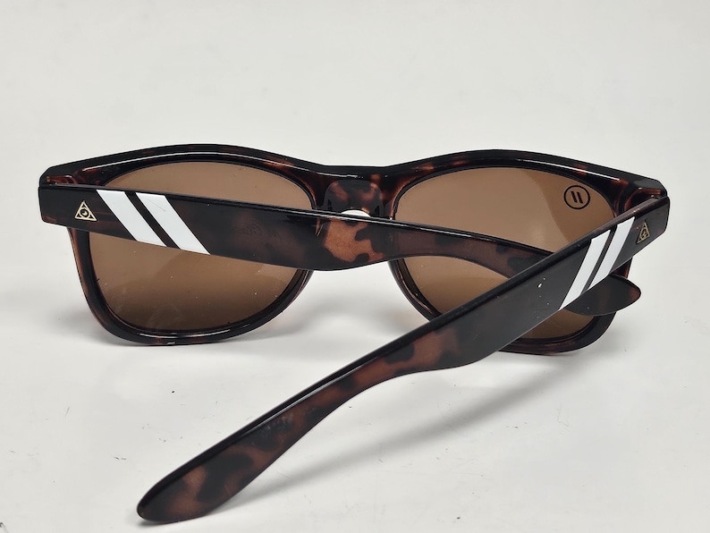 May include: Tortoiseshell sunglasses with brown lenses. The temples feature two white diagonal stripes and a small gold triangular logo near the hinges. A circular logo with two vertical lines is visible on the upper corner of the right lens.