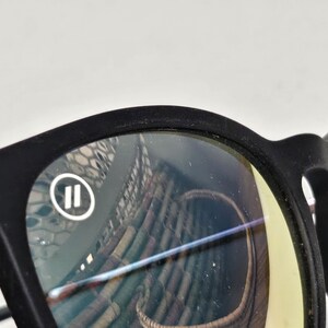 May include: Matte black eyeglasses featuring a rounded frame design. The left lens displays a small white circular logo containing two vertical parallel lines. The lenses have a slight yellow tint and show reflections of a woven basket pattern.