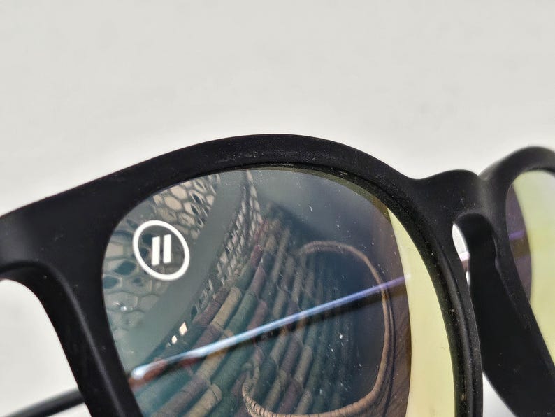 May include: Matte black eyeglasses featuring a rounded frame design. The left lens displays a small white circular logo containing two vertical parallel lines. The lenses have a slight yellow tint and show reflections of a woven basket pattern.