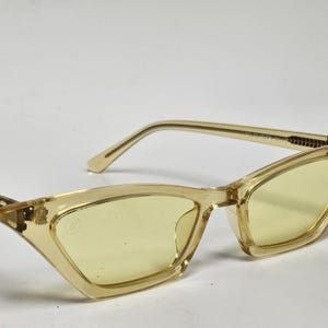 K&ouml;nnte beinhalten: Katzenaugen-Sonnenbrille mit einem durchscheinenden, blassgelben Kunststoffrahmen und passenden hellgelb get&ouml;nten Gl&auml;sern. Die B&uuml;gel haben ein strukturiertes Metalldetail an den Scharnieren und ein kleines kreisf&ouml;rmiges Logo auf dem linken Glas.