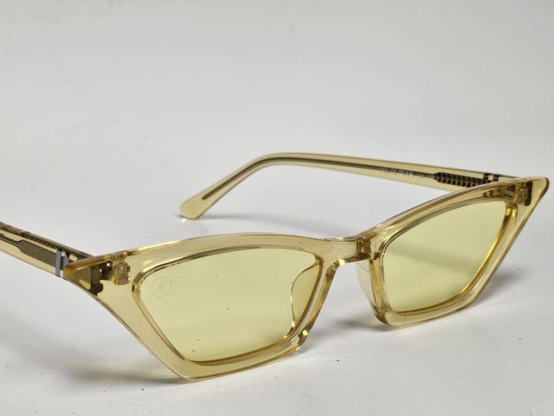 K&ouml;nnte beinhalten: Katzenaugen-Sonnenbrille mit einem durchscheinenden, blassgelben Kunststoffrahmen und passenden hellgelb get&ouml;nten Gl&auml;sern. Die B&uuml;gel haben ein strukturiertes Metalldetail an den Scharnieren und ein kleines kreisf&ouml;rmiges Logo auf dem linken Glas.