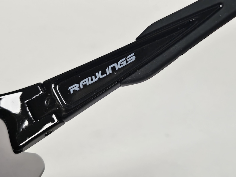 K&ouml;nnte beinhalten: Nahaufnahme eines schwarzen Sportbrillenb&uuml;gels mit dem Markennamen "RAWLINGS" in wei&szlig;er Schrift. Der B&uuml;gel hat eine gl&auml;nzende Oberfl&auml;che und ein strukturiertes Design.