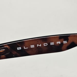 May include: A close-up view of the temple arm of a pair of sunglasses featuring a brown and black tortoiseshell pattern. The word BLENDERS is printed in white capital letters on the side of the arm, which also includes a metallic hinge detail.