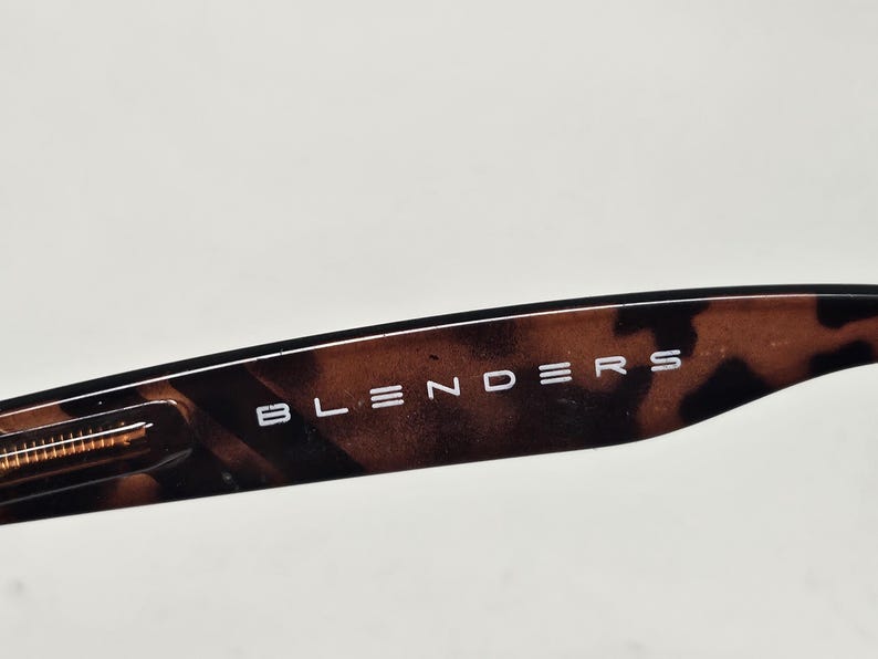 May include: A close-up view of the temple arm of a pair of sunglasses featuring a brown and black tortoiseshell pattern. The word BLENDERS is printed in white capital letters on the side of the arm, which also includes a metallic hinge detail.