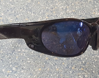 Może przedstawiać: Czarne okulary przeciwsłoneczne z niebieskimi soczewkami. Okulary mają owijany design z grubą oprawką i nowoczesną estetyką. Soczewki wydają się odblaskowe, a oprawka ma matowe wykończenie.