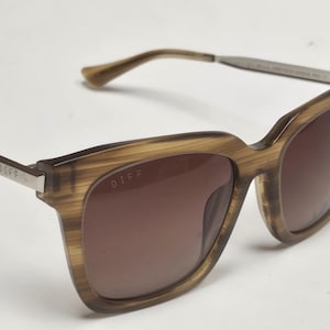Puede incluir: Gafas de sol marrones y plateadas con un patrón de veta de madera en las monturas. El nombre de la marca "DIFF" está impreso en el lateral de las monturas.