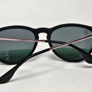 May include: Sunglasses with a matte black frame and dark grey lenses. The temples feature a metallic rose gold finish with thin silver accents near the hinges. A small circular logo with two vertical lines is visible on the upper right lens.