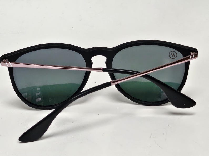 May include: Sunglasses with a matte black frame and dark grey lenses. The temples feature a metallic rose gold finish with thin silver accents near the hinges. A small circular logo with two vertical lines is visible on the upper right lens.