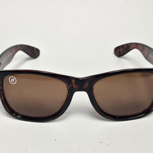 May include: Tortoiseshell-patterned sunglasses with brown-tinted lenses. The frames feature a dark brown and black mottled design. A small white circular logo with two vertical lines is printed on the upper corner of the left lens.