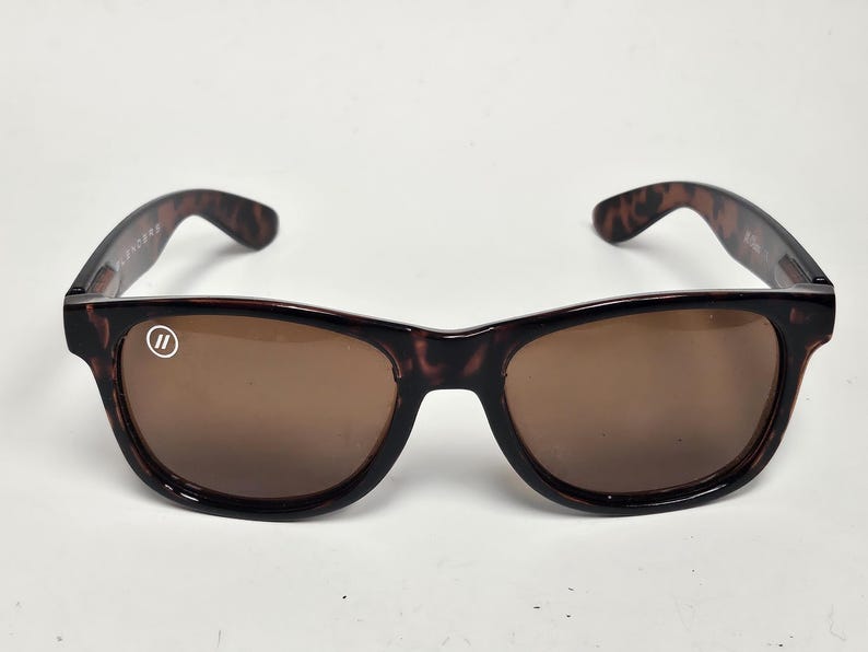 May include: Tortoiseshell-patterned sunglasses with brown-tinted lenses. The frames feature a dark brown and black mottled design. A small white circular logo with two vertical lines is printed on the upper corner of the left lens.