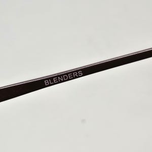 May include: A dark brown eyeglass temple arm featuring the word BLENDERS printed in white capital letters. The arm is made of a smooth, solid material and includes a small metal hinge component at the far left end of the frame structure.