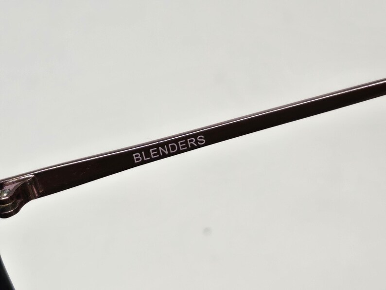May include: A dark brown eyeglass temple arm featuring the word BLENDERS printed in white capital letters. The arm is made of a smooth, solid material and includes a small metal hinge component at the far left end of the frame structure.