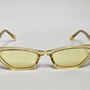 K&ouml;nnte beinhalten: Katzenaugen-Sonnenbrille mit einem durchscheinenden, hellgelben Kunststoffrahmen und passenden gelb get&ouml;nten Gl&auml;sern. Das Design umfasst d&uuml;nne, kantige B&uuml;gel und ein kleines kreisf&ouml;rmiges Logo-Detail an der oberen Ecke des linken Glases.