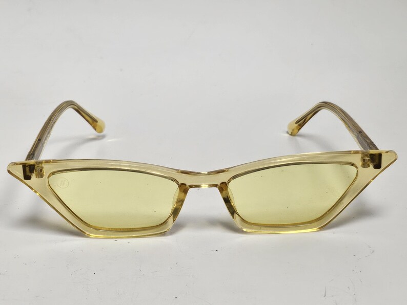 K&ouml;nnte beinhalten: Katzenaugen-Sonnenbrille mit einem durchscheinenden, hellgelben Kunststoffrahmen und passenden gelb get&ouml;nten Gl&auml;sern. Das Design umfasst d&uuml;nne, kantige B&uuml;gel und ein kleines kreisf&ouml;rmiges Logo-Detail an der oberen Ecke des linken Glases.