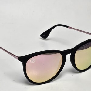 May include: Sunglasses with a matte black frame and thin metallic rose gold temples. The lenses feature a reflective pink and gold gradient finish. A small circular logo with two vertical lines is visible on the upper corner of the left lens.
