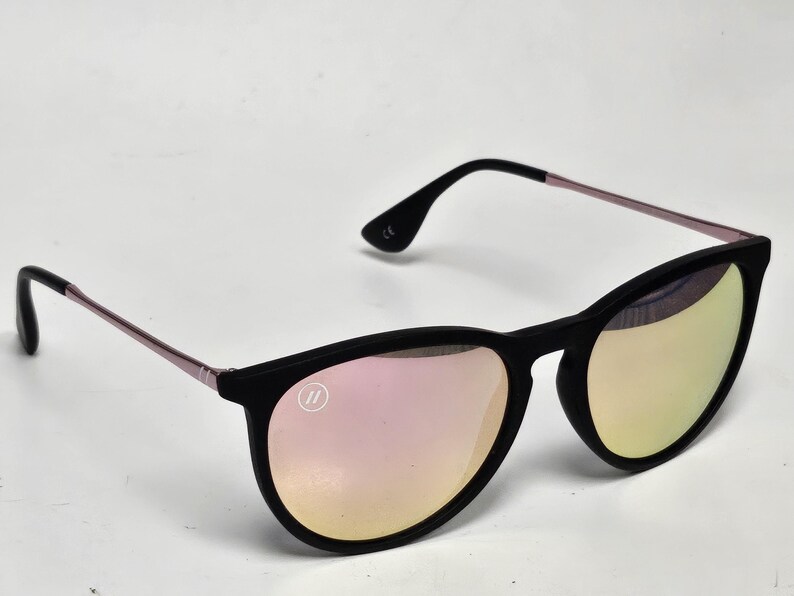 May include: Sunglasses with a matte black frame and thin metallic rose gold temples. The lenses feature a reflective pink and gold gradient finish. A small circular logo with two vertical lines is visible on the upper corner of the left lens.