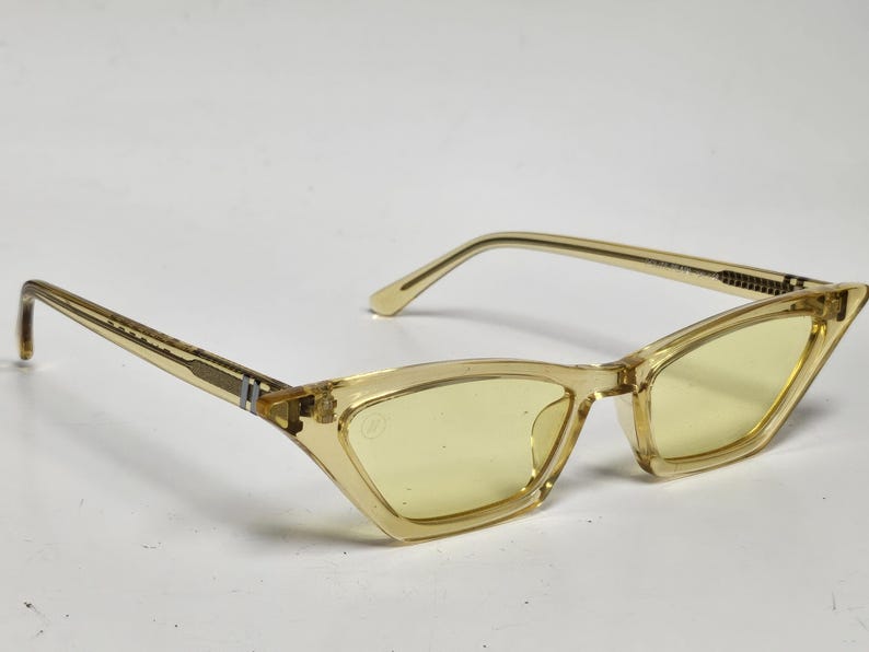 K&ouml;nnte beinhalten: Katzenaugenbrille mit einem durchscheinenden hellgelben Rahmen und passenden hellgelb get&ouml;nten Gl&auml;sern. Die B&uuml;gel sind mit silberfarbenen Metallscharnieren am Rahmen befestigt und weisen ein kleines rechteckiges Detail in Silberoptik auf.