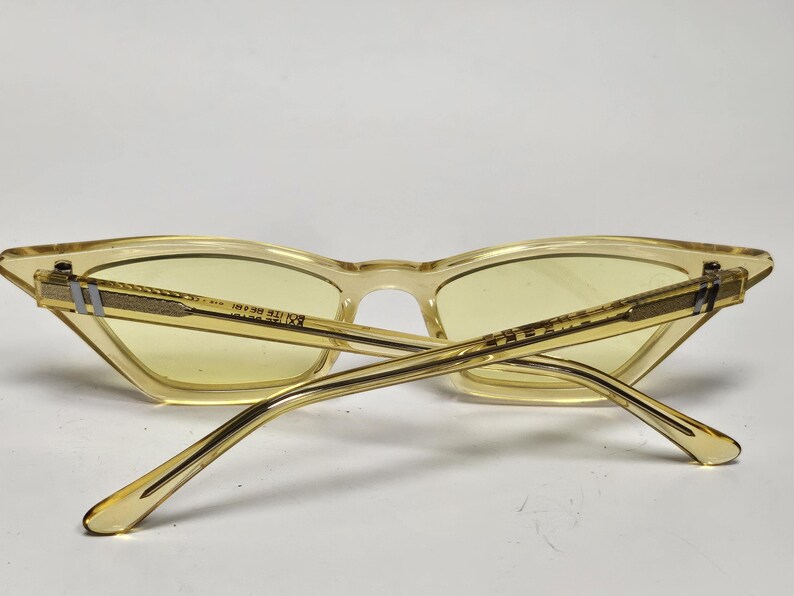 K&ouml;nnte beinhalten: Katzenaugen-Sonnenbrille mit einem durchscheinenden blassgelben Rahmen und passenden hellgelb get&ouml;nten Gl&auml;sern. Die B&uuml;gel verf&uuml;gen &uuml;ber eine strukturierte goldfarbene Metalleinlage mit einem kleinen silberfarbenen Akzent am Scharnier.
