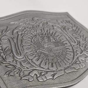 Op de afbeelding: Zilverkleurige, schildvormige gesp met een reliëf ontwerp. Het ontwerp omvat een adelaar, zonnestralen en tekst, omgeven door lauriertakken. De gesp heeft een getextureerd oppervlak en een gebogen vorm.